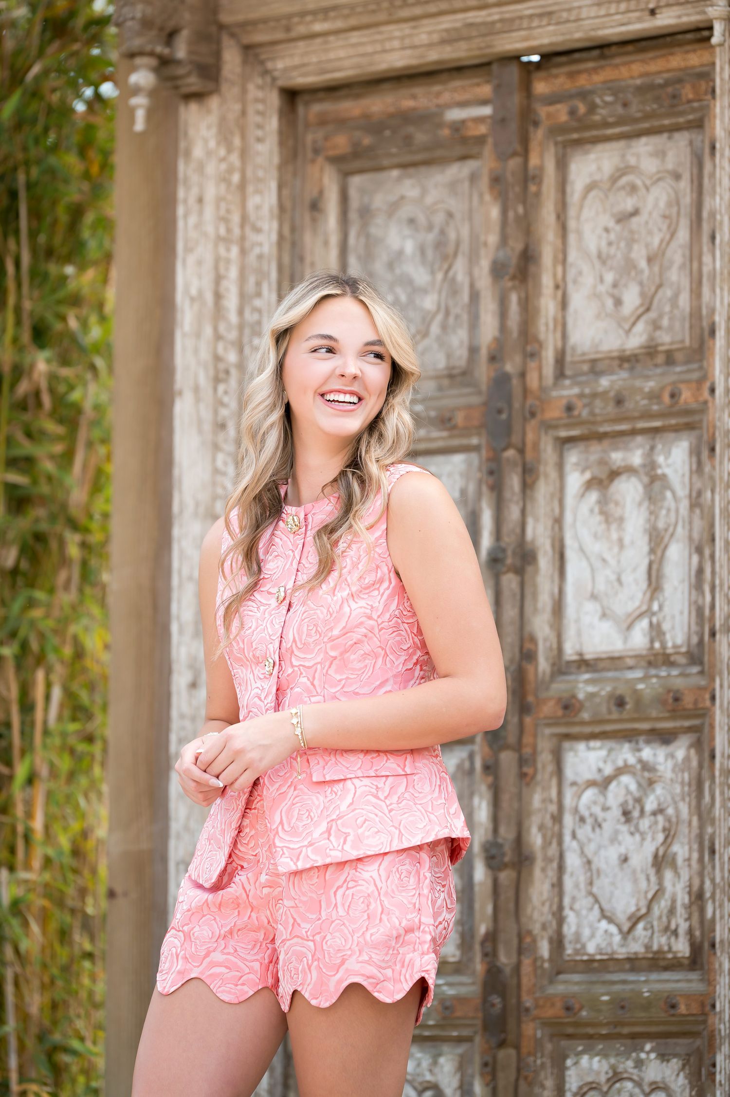 Smiling young woman in pink lace top and shorts standing before ornate vintage wooden doors outdoors.