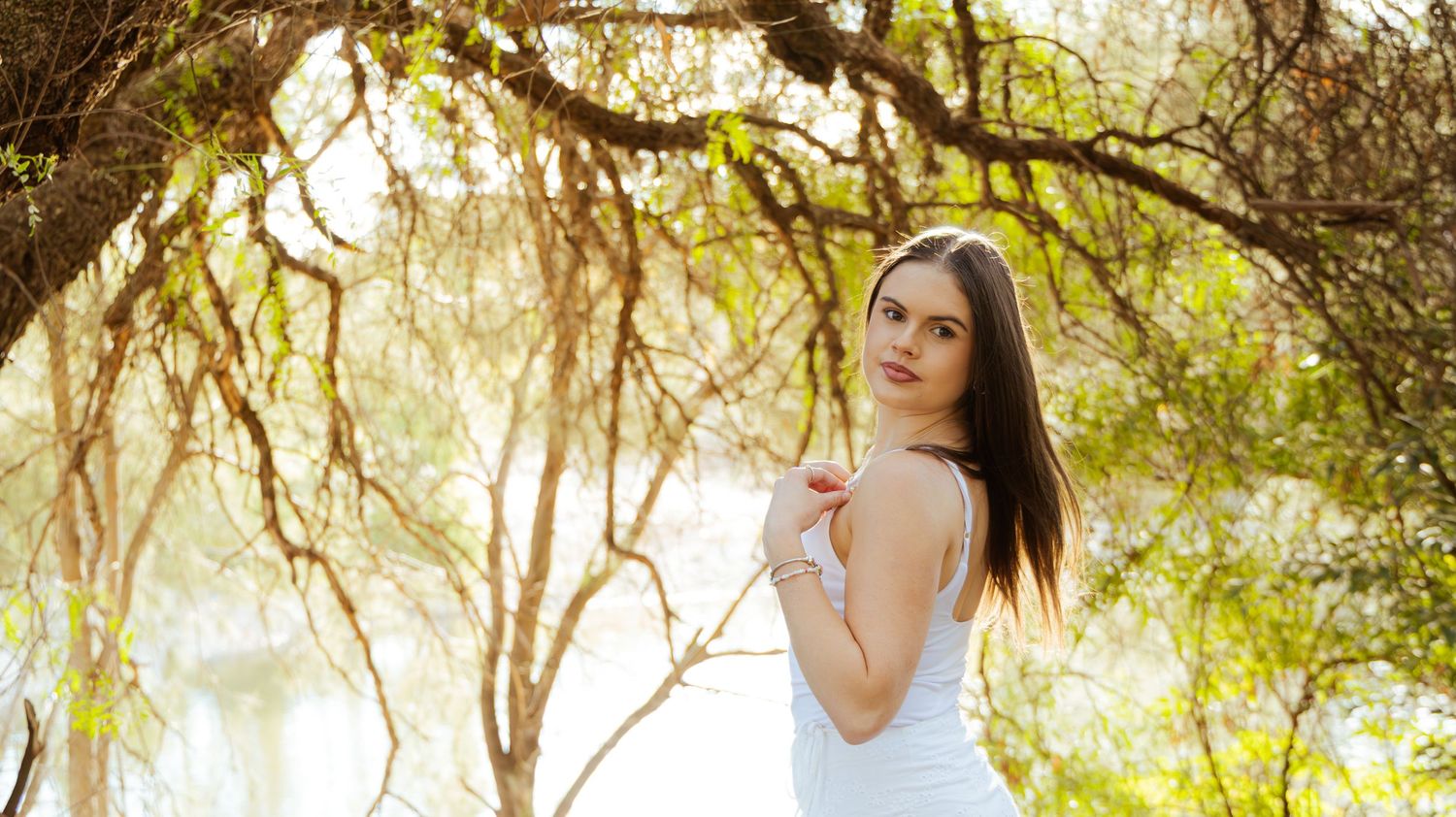 Young woman in white dress posing gracefully under moss-covered tree branches in soft sunlight.