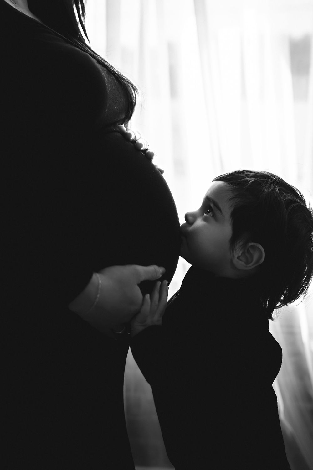 Black and white photo of a young child kissing pregnant mother's belly in a tender maternity moment.
