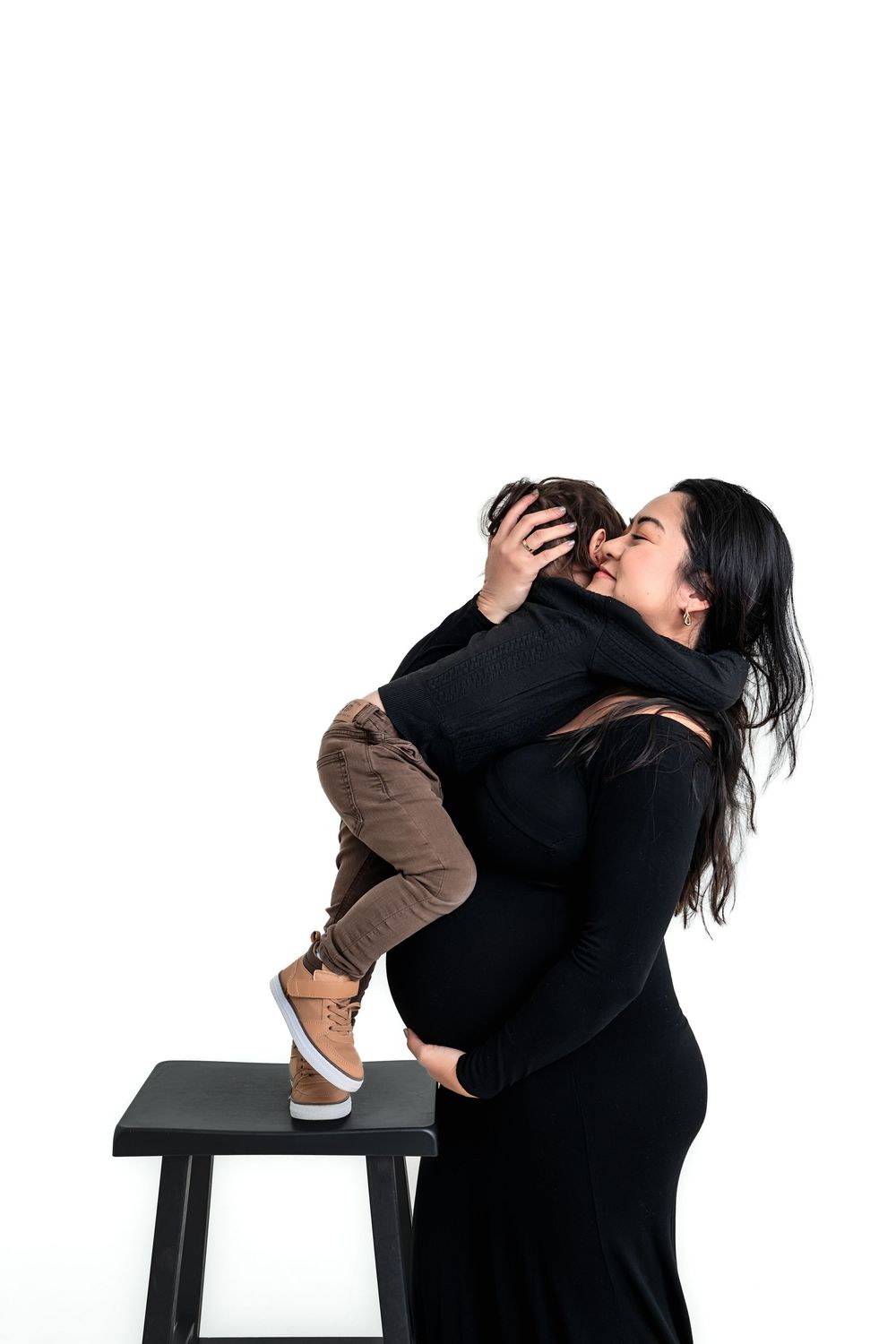 Pregnant woman in black dress holding and kissing her toddler son in a studio setting.