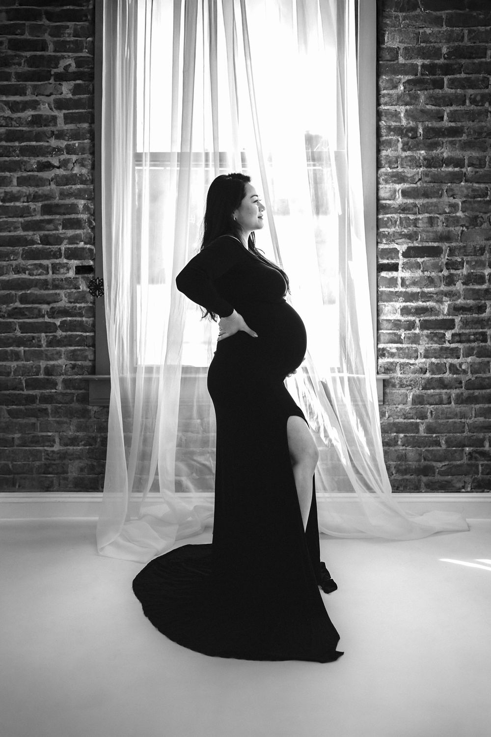 Black and white maternity photo of pregnant woman in elegant black gown standing by window with sheer curtains.