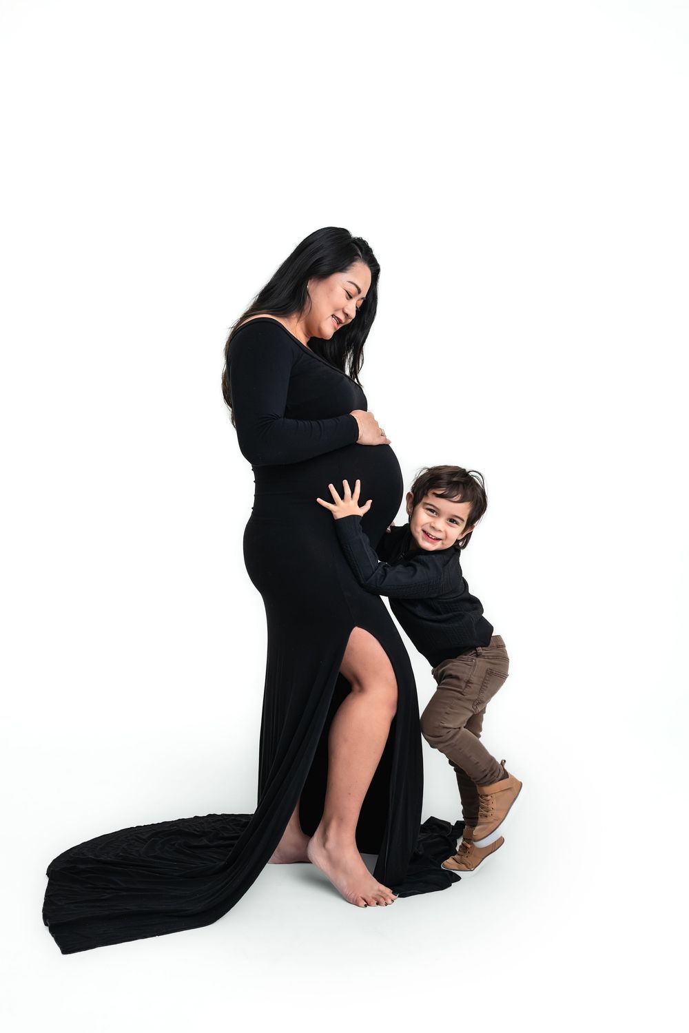 Pregnant woman in elegant black maternity gown smiling with her young son hugging her baby bump against a pure white backdrop.