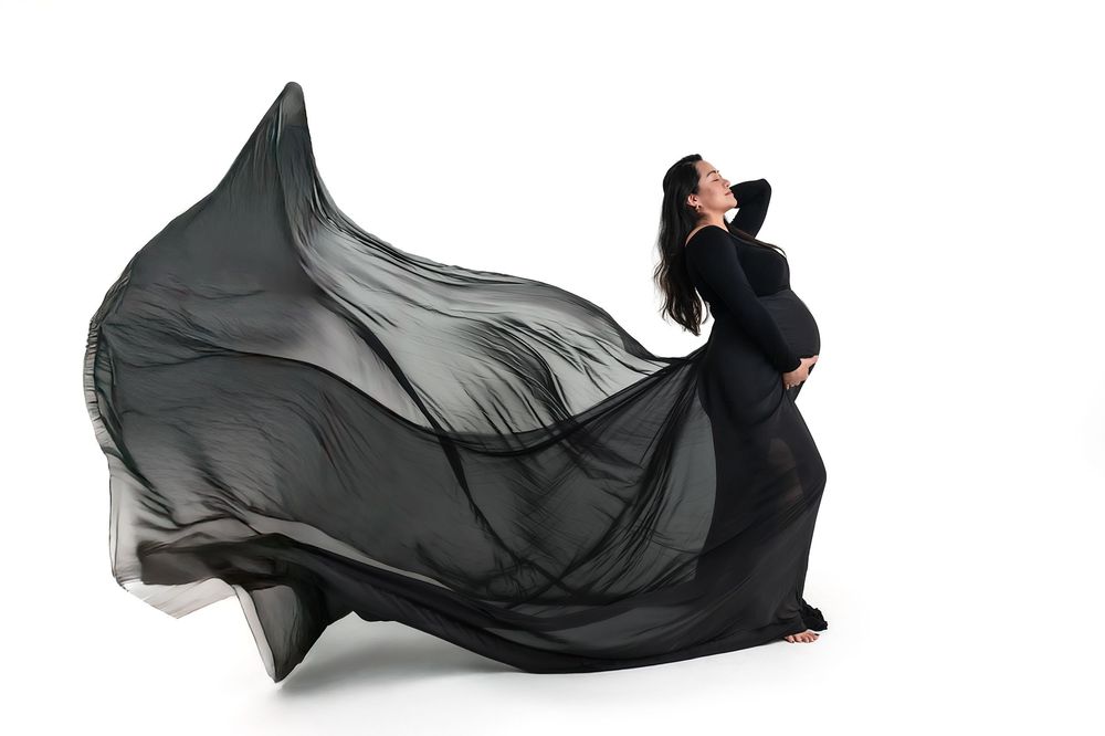 Pregnant woman in flowing black maternity gown with dramatic fabric billowing against white background.