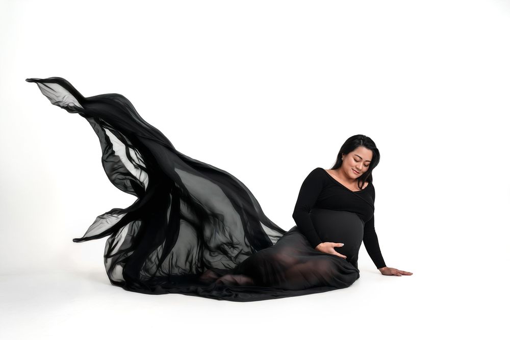 Pregnant woman in black flowing gown posed gracefully on floor in studio setting with white backdrop.