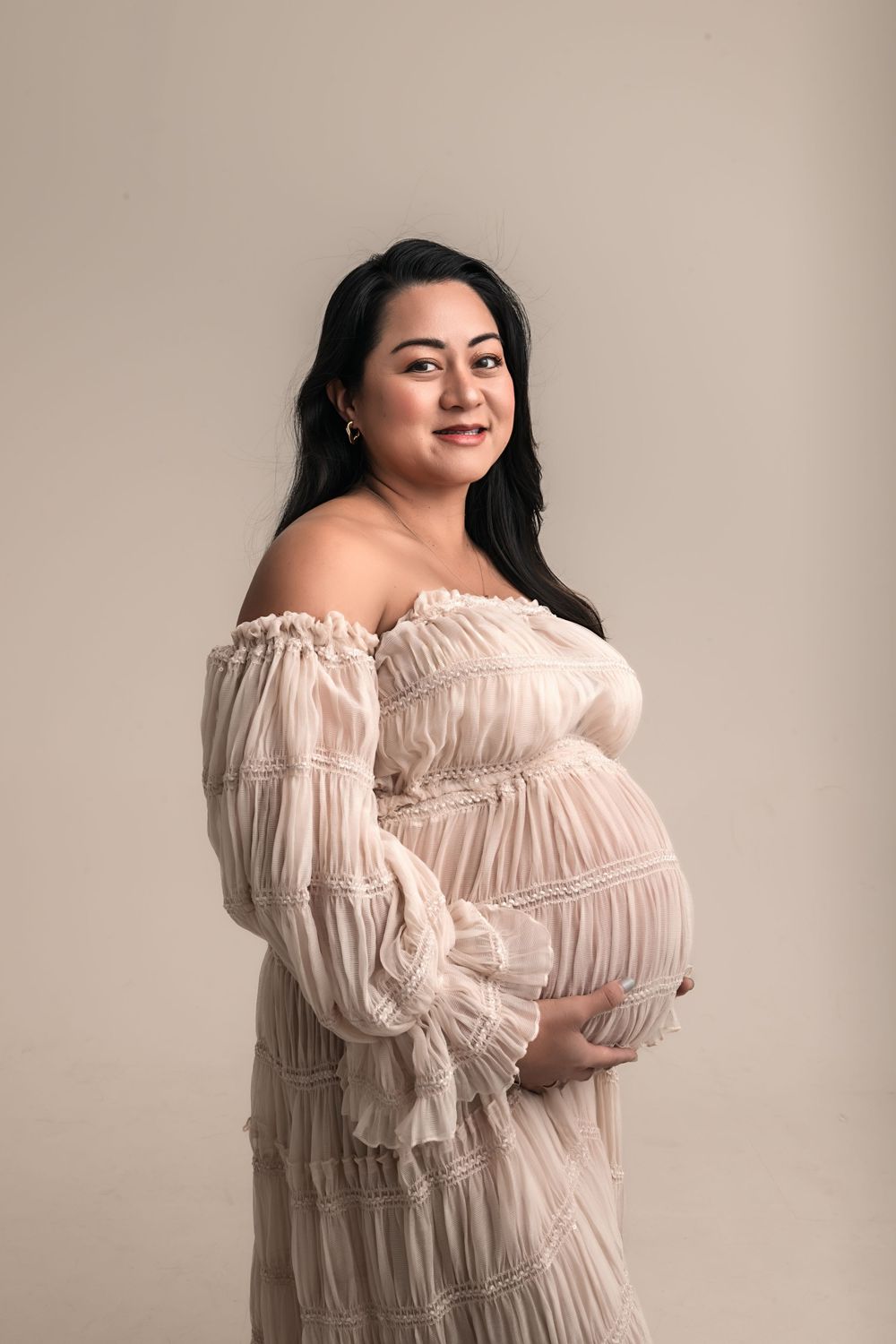 Smiling pregnant woman in off-shoulder cream ruffled dress holding baby bump for studio maternity photos in Marietta.