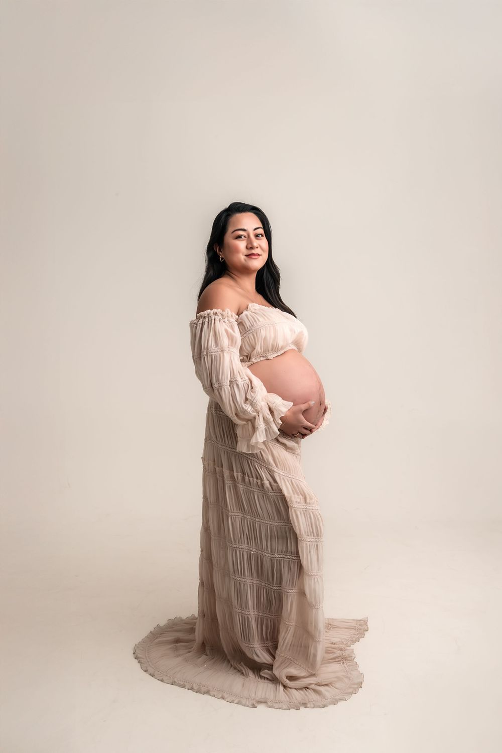 Pregnant woman in sheer blush off-shoulder gown cradling baby bump in elegant studio maternity portrait.