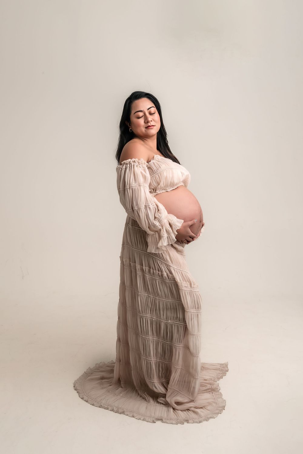 Pregnant woman in flowing off-shoulder blush dress cradling baby bump in soft studio maternity portrait.