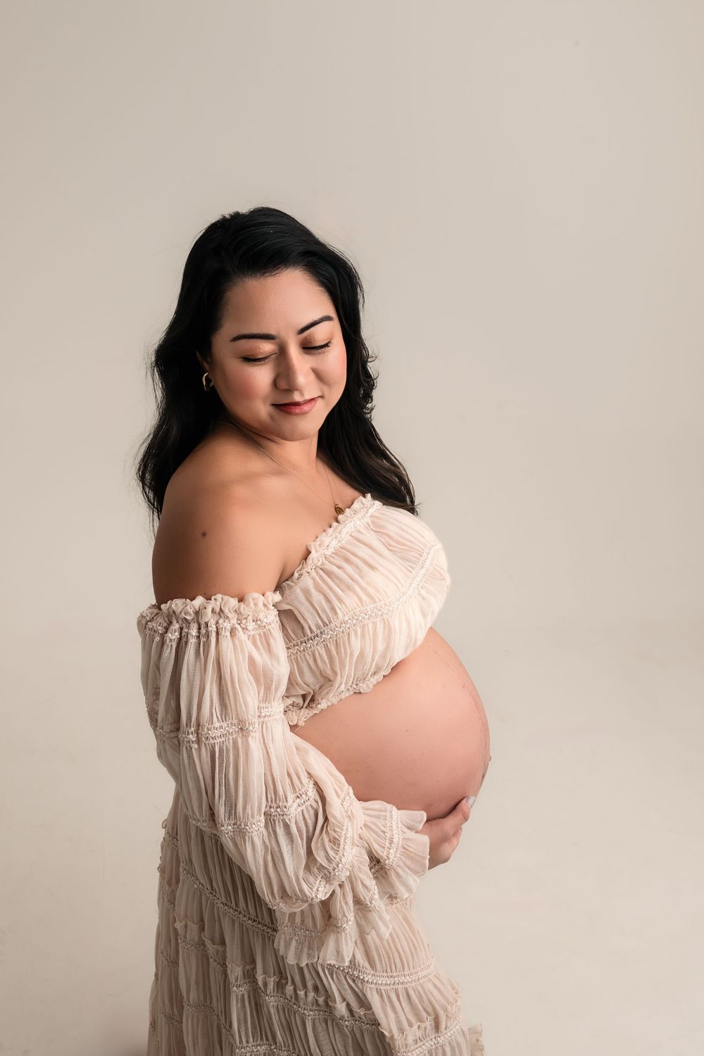 Pregnant woman in off-shoulder beige lace dress cradling her bare baby bump in a studio for Atlanta maternity photographer.