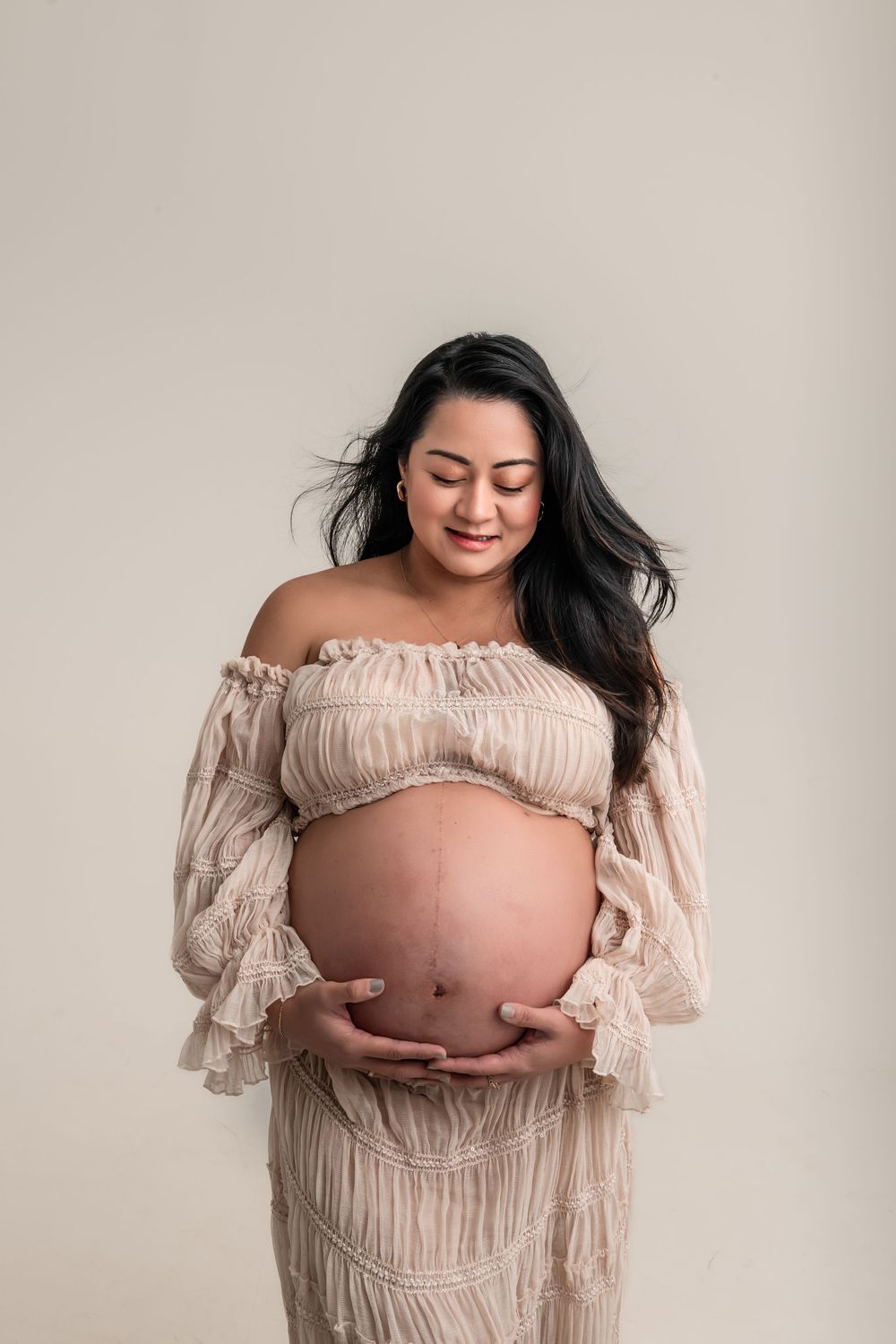 Smiling pregnant woman in boho blush two-piece dress gently holding bare baby bump in studio portrait.