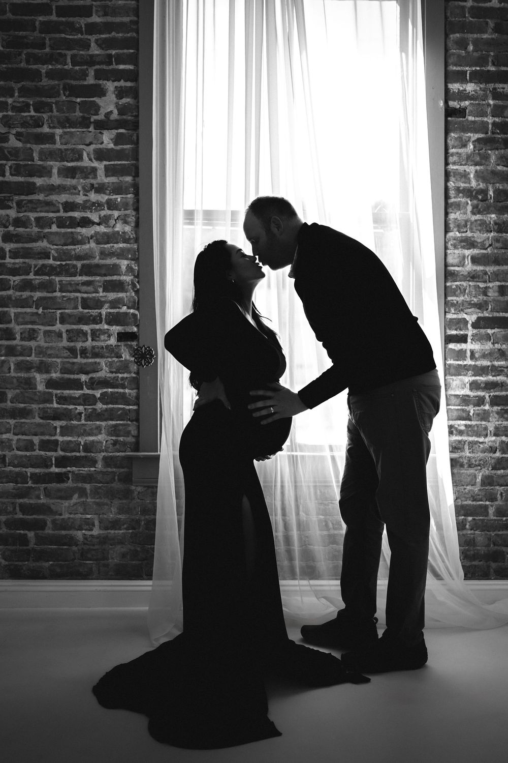 Black and white silhouette of pregnant couple kissing by window in a studio with brick walls.