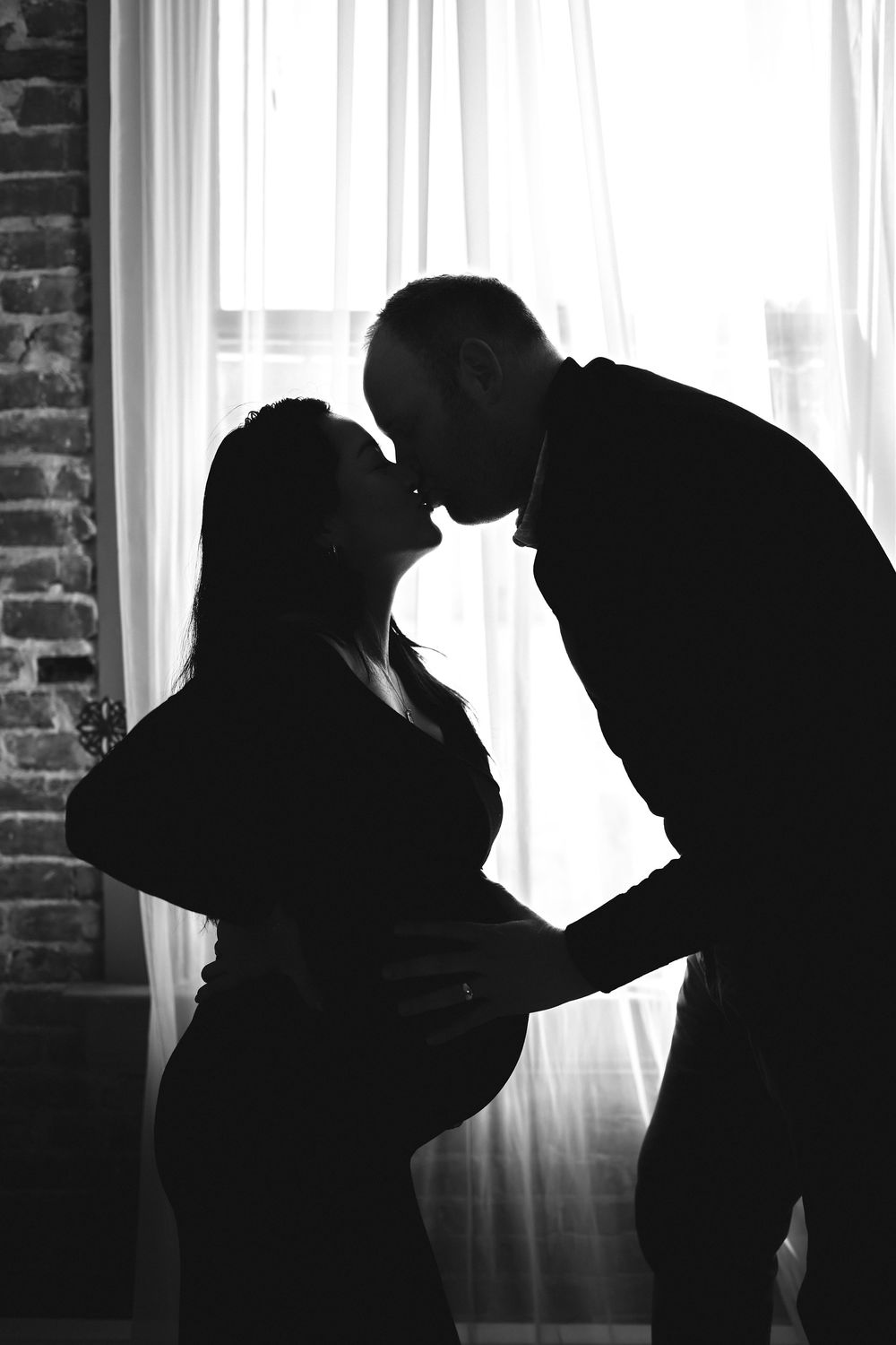Black and white silhouette of a couple kissing in front of a bright window during a maternity photo session.