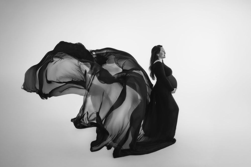 Black and white maternity photo of pregnant woman in flowing black gown with fabric billowing dramatically in studio.