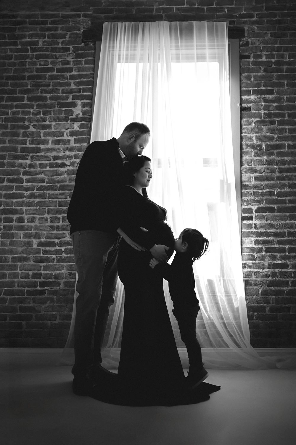 Black and white silhouette of a pregnant mom with father and child by a brick wall and bright window.