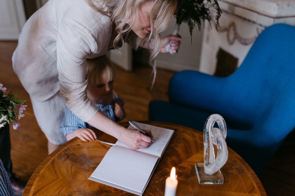 Woman signing a guest book at a wedding while a young child watches nearby. Borgerlig vigsel i Karlskrona på Nordenskjöldska gården.