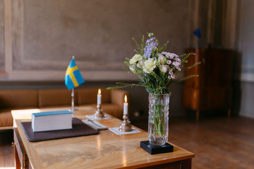 A wooden table with a Swedish flag, lit candle, books, and a glass vase with flowers. Borgerlig vigsel i Karlskrona på Nordenskjöldska gårde