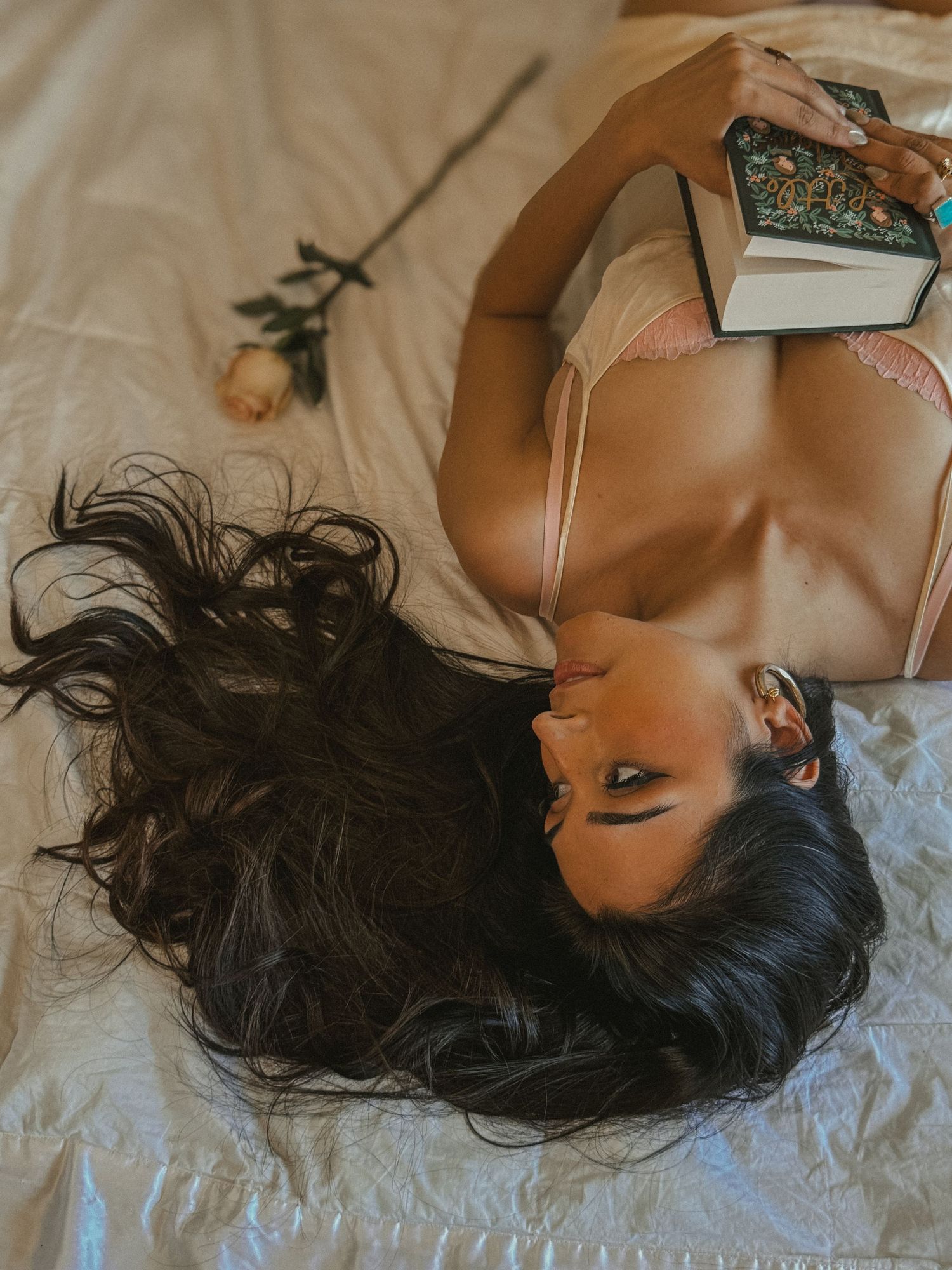 Woman with long dark hair lying on bed holding decorative book with a rose beside her.
