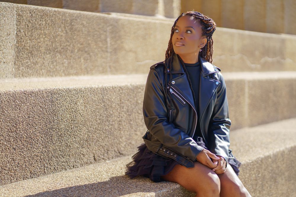 Young Black woman with braids in leather jacket and skirt sitting on stone steps outdoors in sunlight.