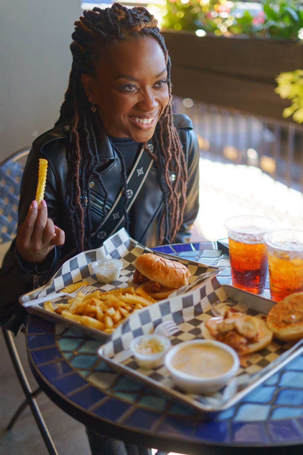 Smiling woman with locs in leather jacket enjoying fries and burger platter with drinks at outdoor cafe table.