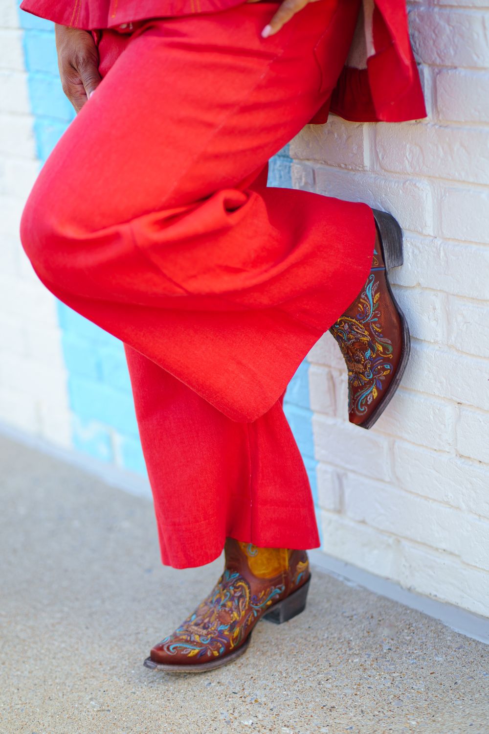 Person wearing vibrant red wide-leg pants and colorful embroidered brown cowboy boots leaning against a white brick wall.