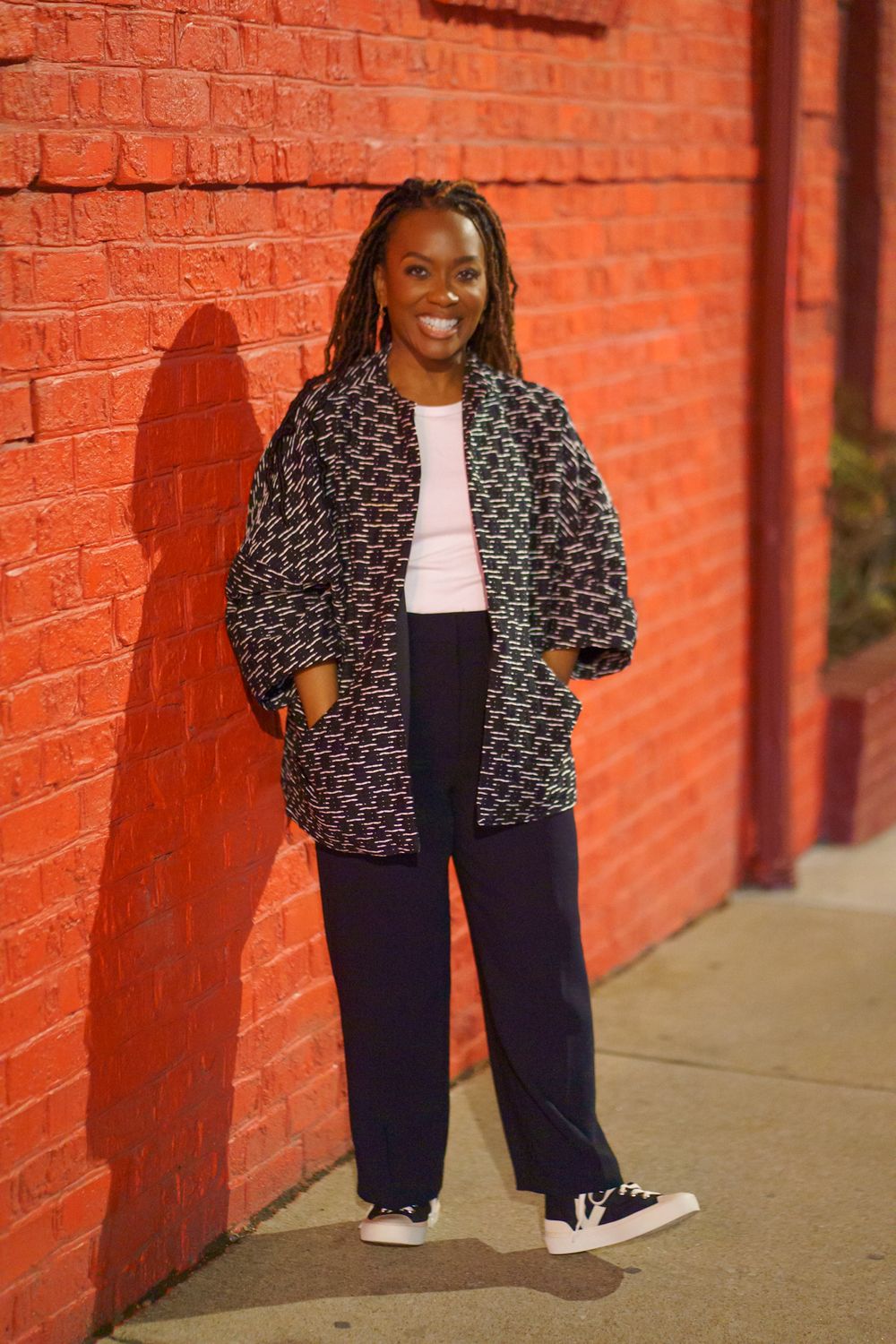Smiling woman in patterned jacket and navy pants leaning against a red brick wall outdoors.