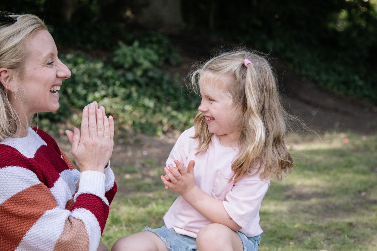 A smiling mother and young blonde girl clapping hands together outdoors in a green park setting.