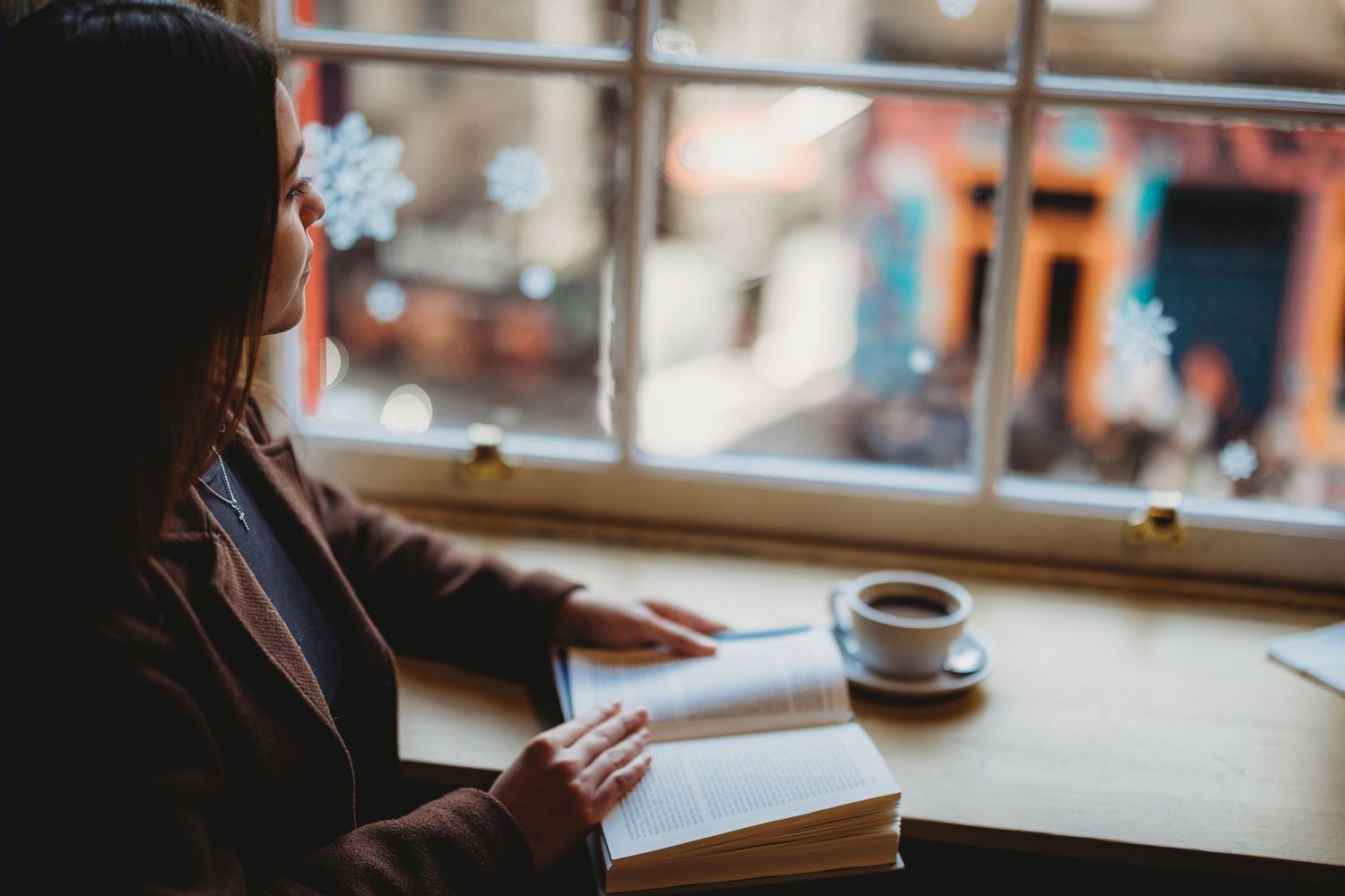 Woman reading a book at a cafe table by a window with a cup of coffee, enjoying a quiet moment indoors.