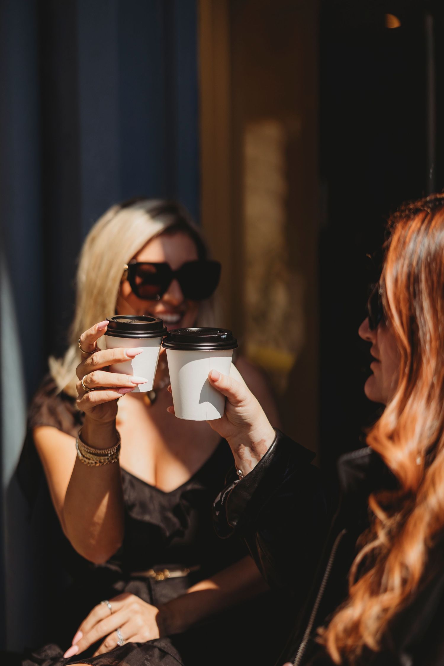 Two stylish women in sunglasses toasting with takeaway coffee cups outdoors in warm sunlight.