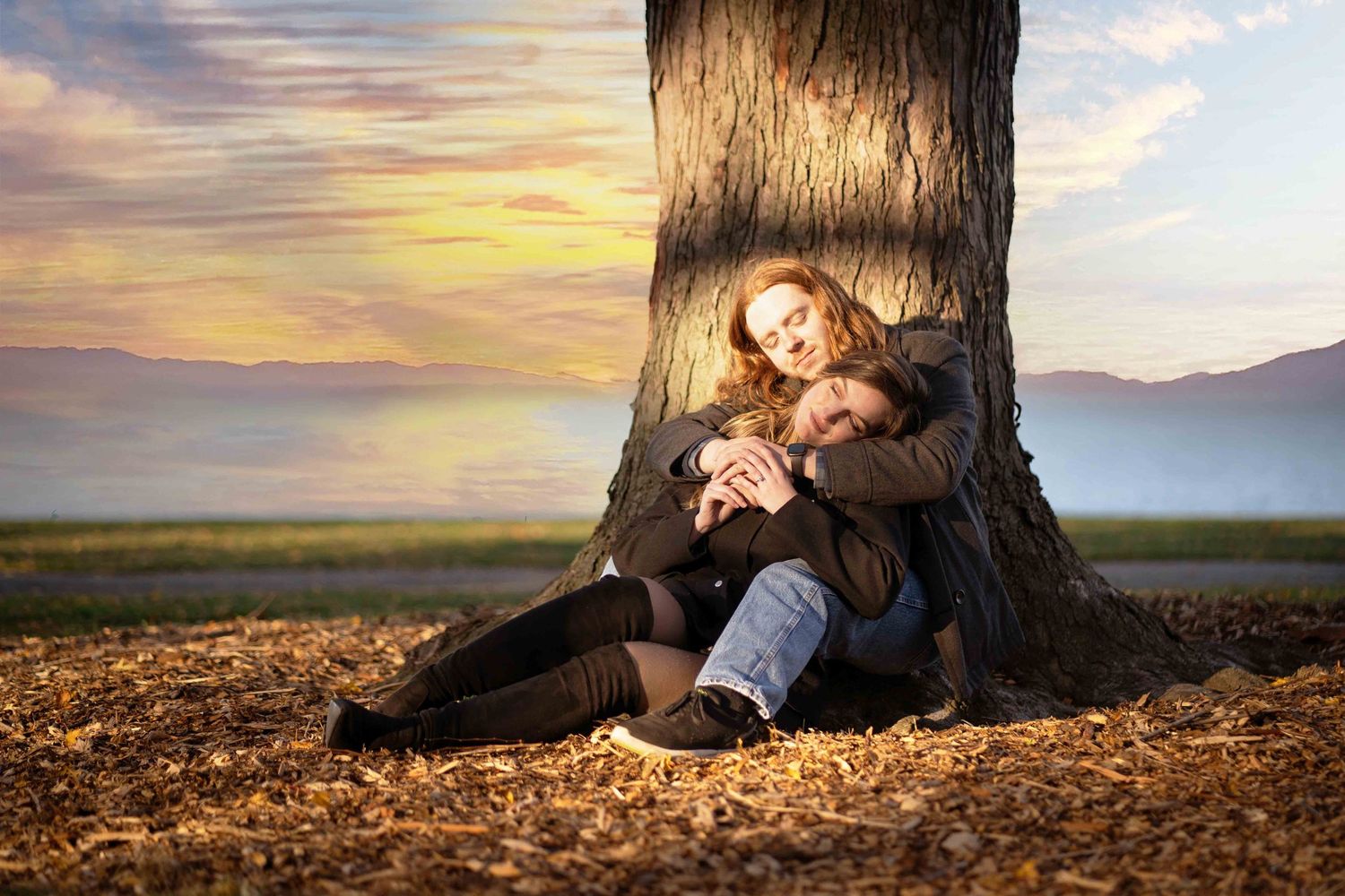 Romantic couple sitting together against a large tree trunk at sunset with colorful sky and autumn leaves.