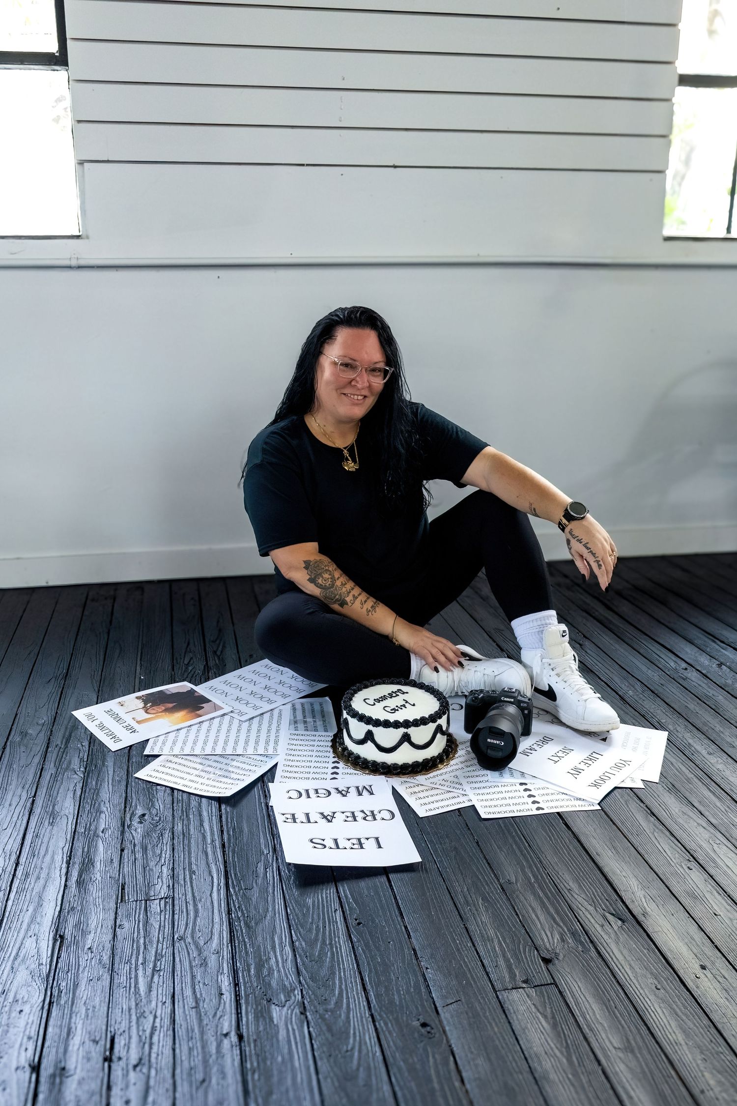 Person in black outfit sitting on wooden floor with a cake, camera, and signs reading Let's Create Magic.