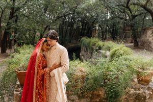 Couple in traditional South Asian wedding attire embracing outdoors, bride in red dupatta and groom in cream sherwani.