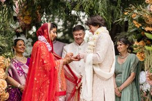 Bride in red sari and groom in ivory sherwani exchange rings during an outdoor Indian wedding ceremony.