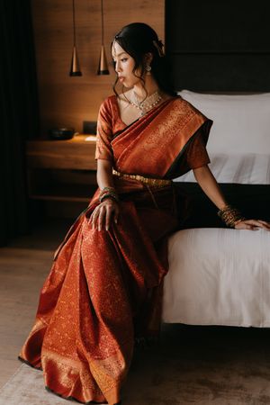Woman in elegant red and gold silk saree with traditional jewelry sits gracefully on bed edge in warmly lit room.