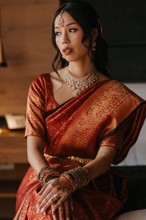 Woman in red silk saree with gold embroidery, wearing traditional Indian bridal jewelry, bangles, and mehndi, seated indoors.