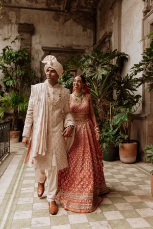 Couple in traditional Indian wedding attire walking through a columned courtyard with potted plants.