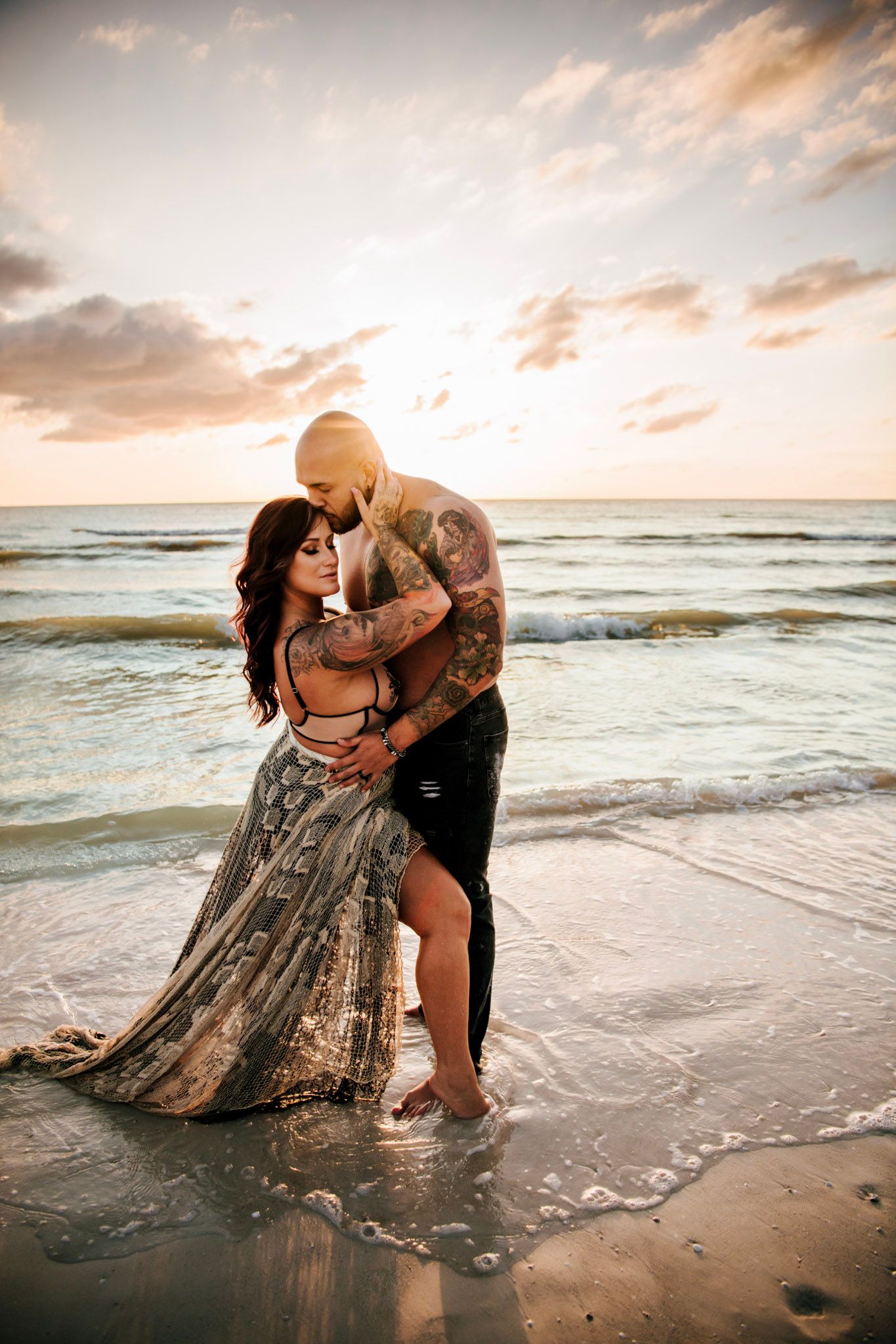 Tattooed couple shares a romantic embrace in shallow water during a beach photography session. The sun is golden and reflects on the water.