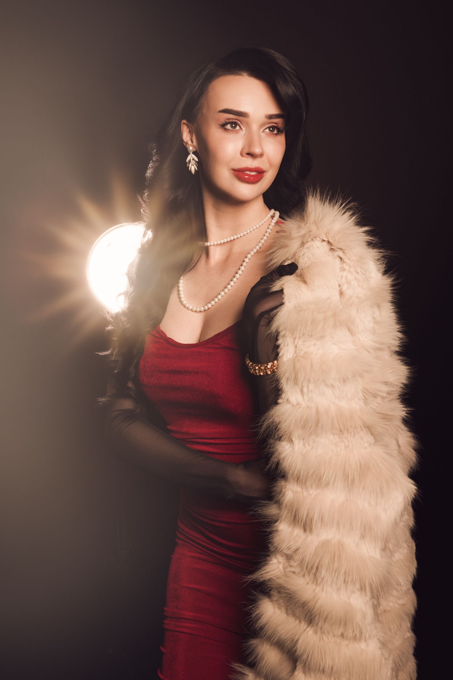 Elegant woman in red dress with pearl necklace, black gloves, and white feather boa posing in dramatic studio lighting.