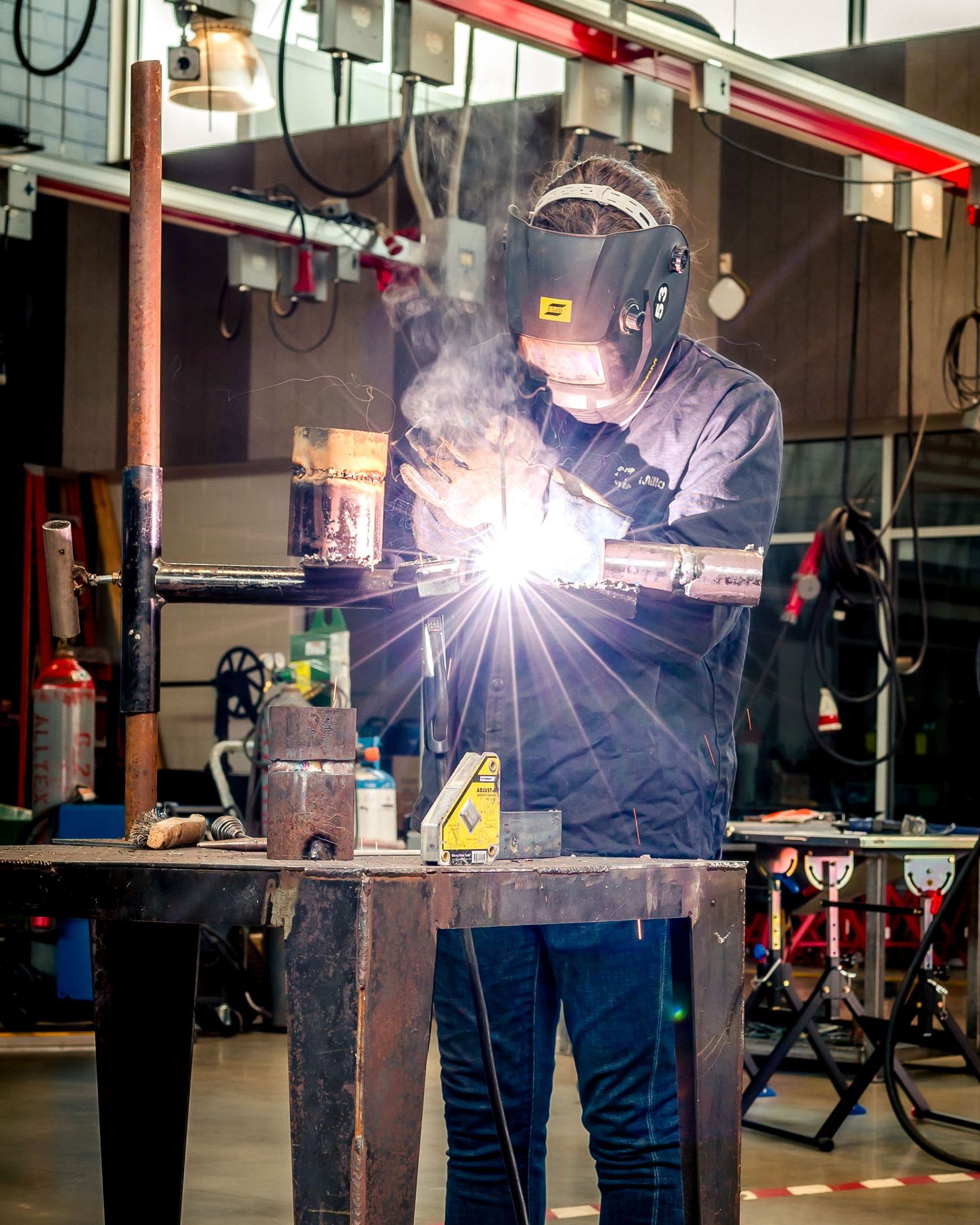 A welder in protective gear and helmet works with bright sparks flying in an industrial workshop setting.