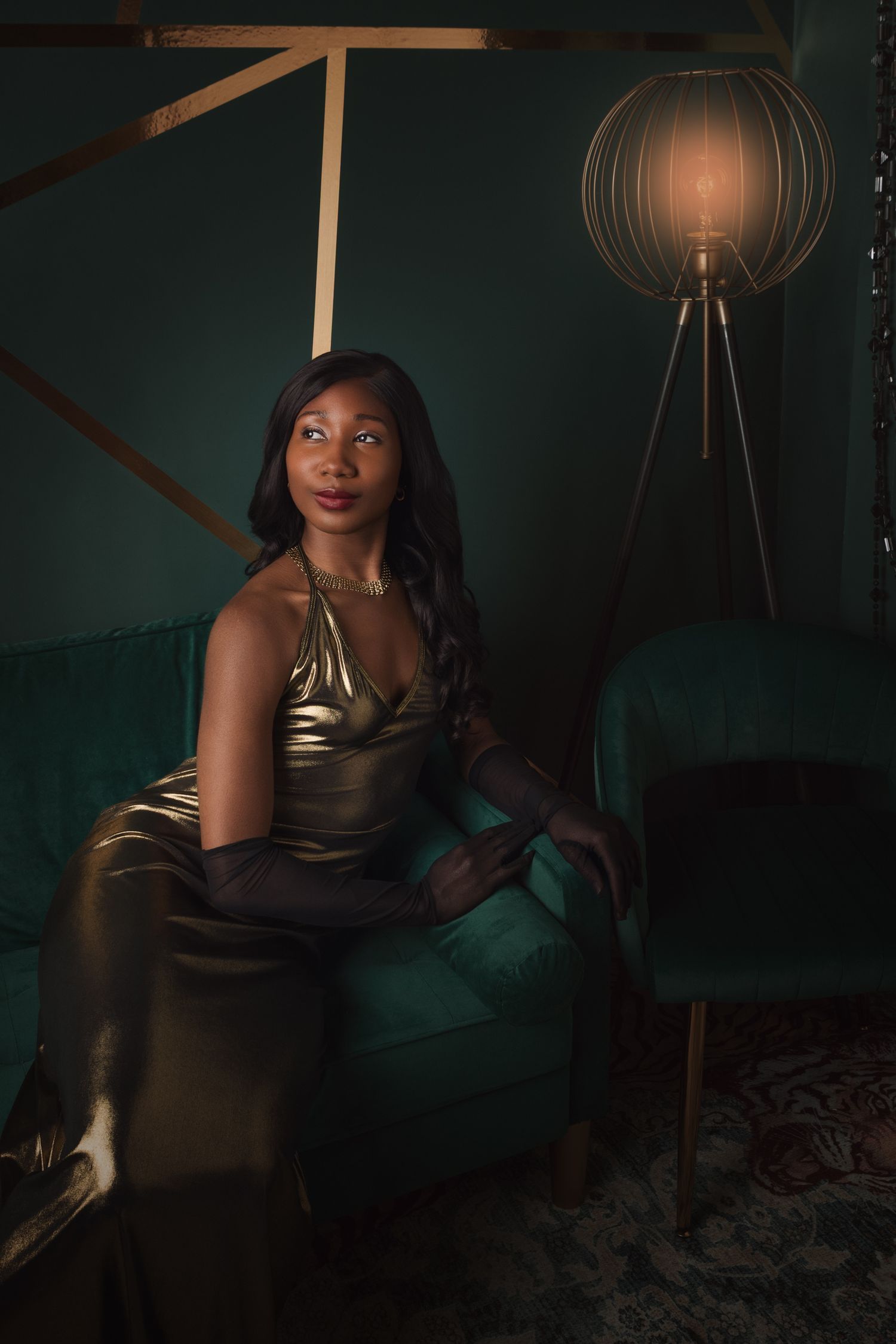 Elegant Black woman in gold satin gown and long gloves seated on green velvet chair in moody, dark interior.