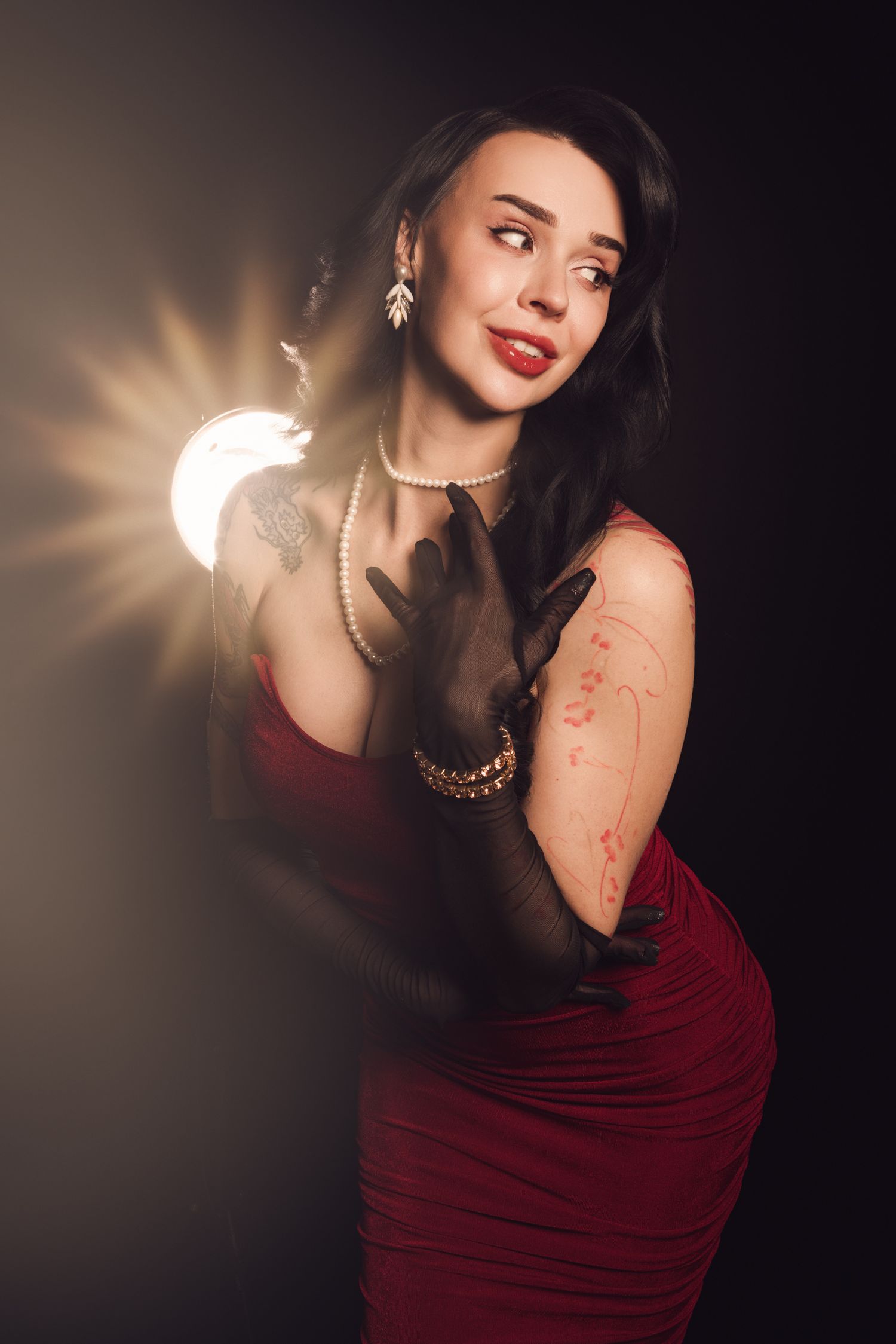 Elegant woman in red dress with black gloves, pearl necklace, and tattoos posing dramatically against dark background with bright light.