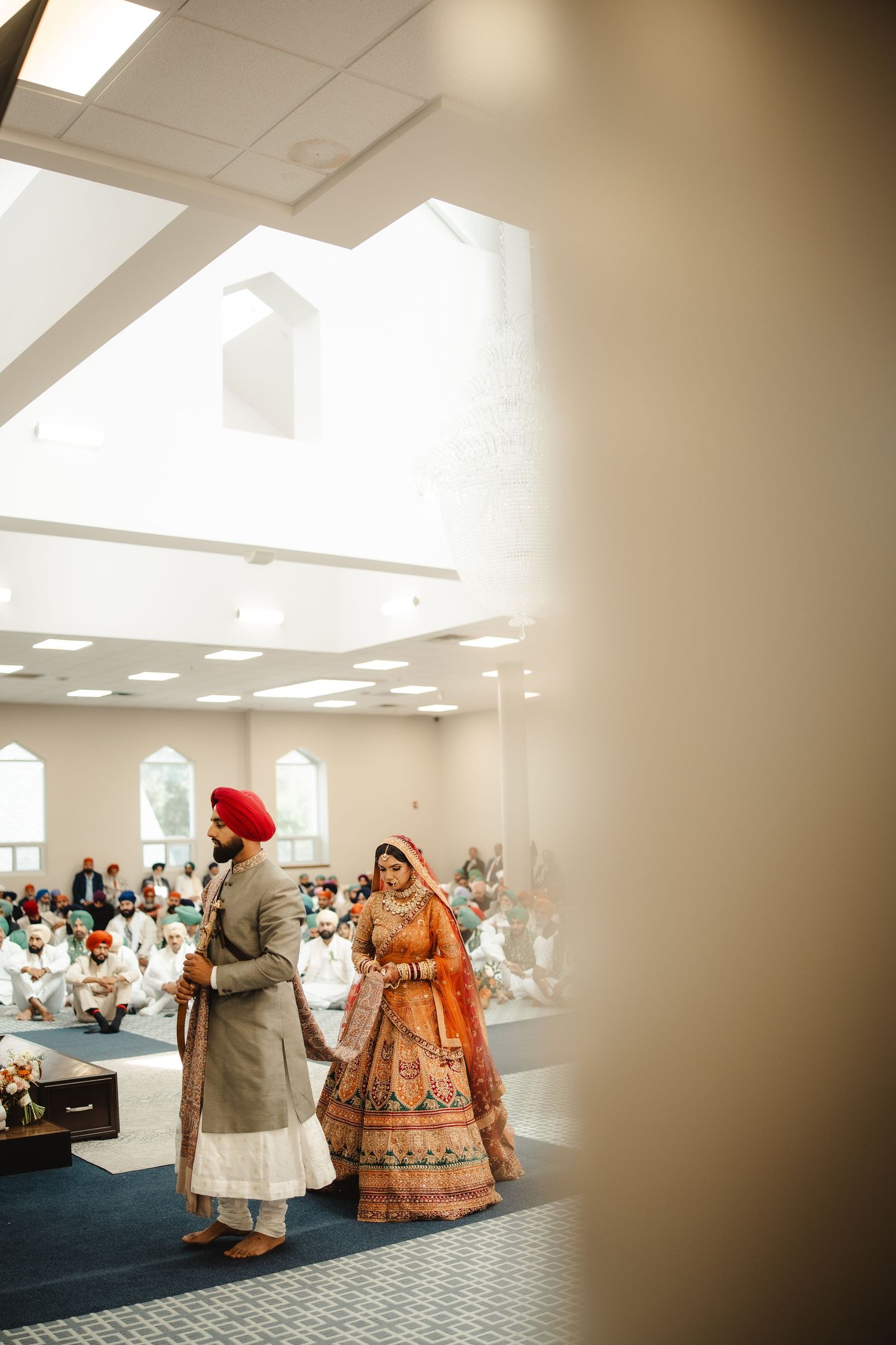 Sikh bride in orange lehenga and groom in red turban walk together during Anand Karaj wedding ceremony.