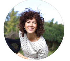 Smiling woman with curly brown hair wearing a polka dot top, posing outdoors with green trees in background.
