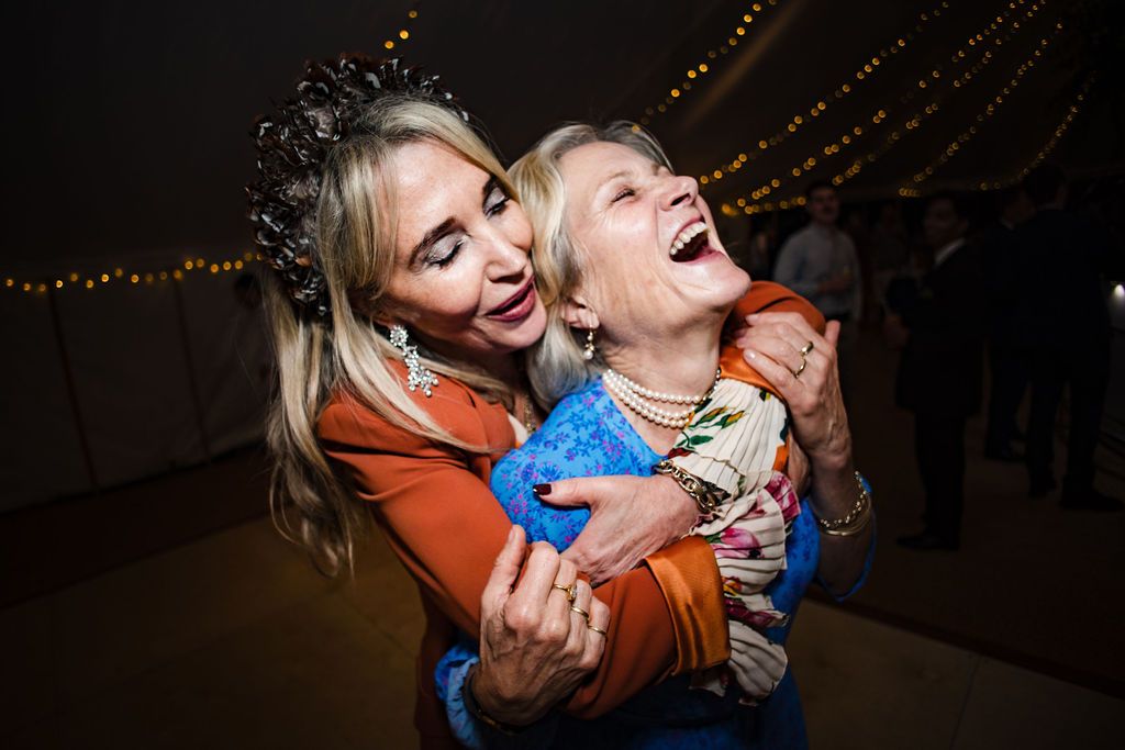 Two women laughing and embracing joyfully at a festive evening event with warm string lights in background.