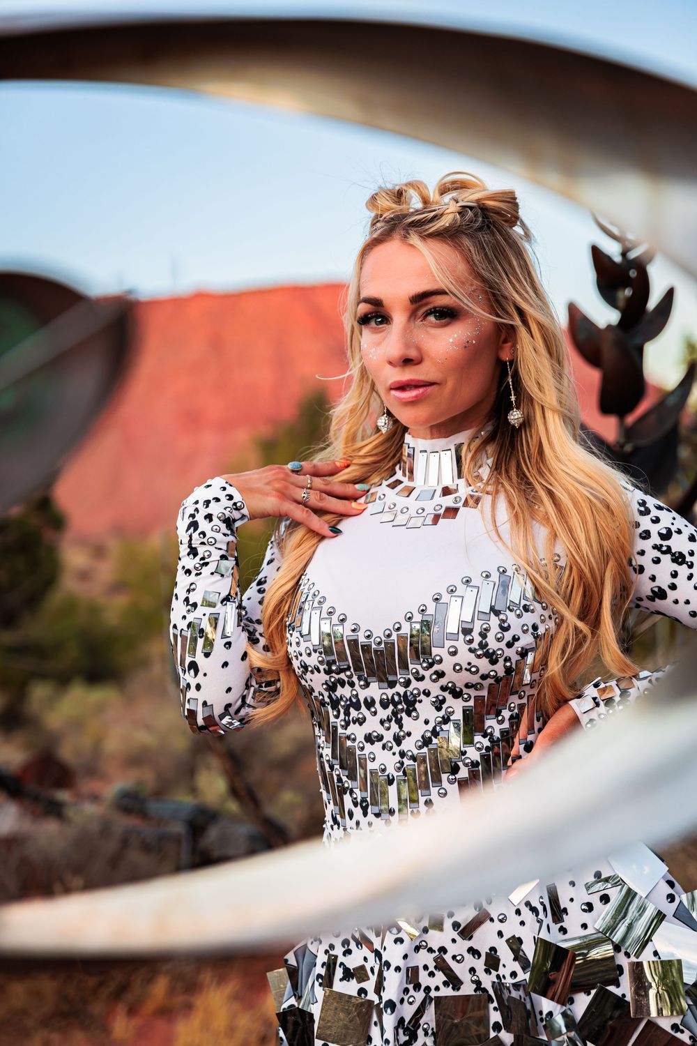 Blonde woman in a sparkling white mirror-embellished dress posing outdoors through a circular metal frame.