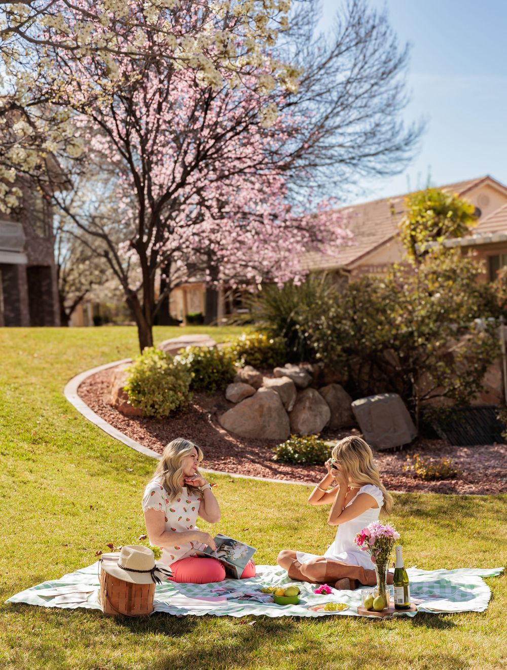 Two women enjoy a spring picnic on a blanket in a lush garden with blooming pink cherry blossom trees.
