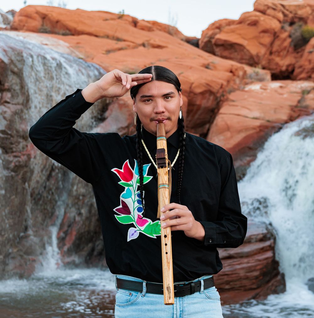 Young Native American man playing a wooden flute near a waterfall, wearing a black floral shirt.