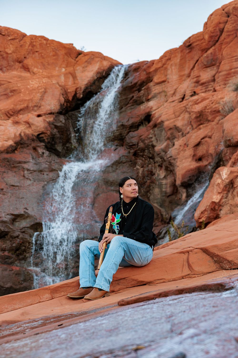 Man sitting on red rock sandstone formations before a cascading waterfall in a desert canyon landscape.