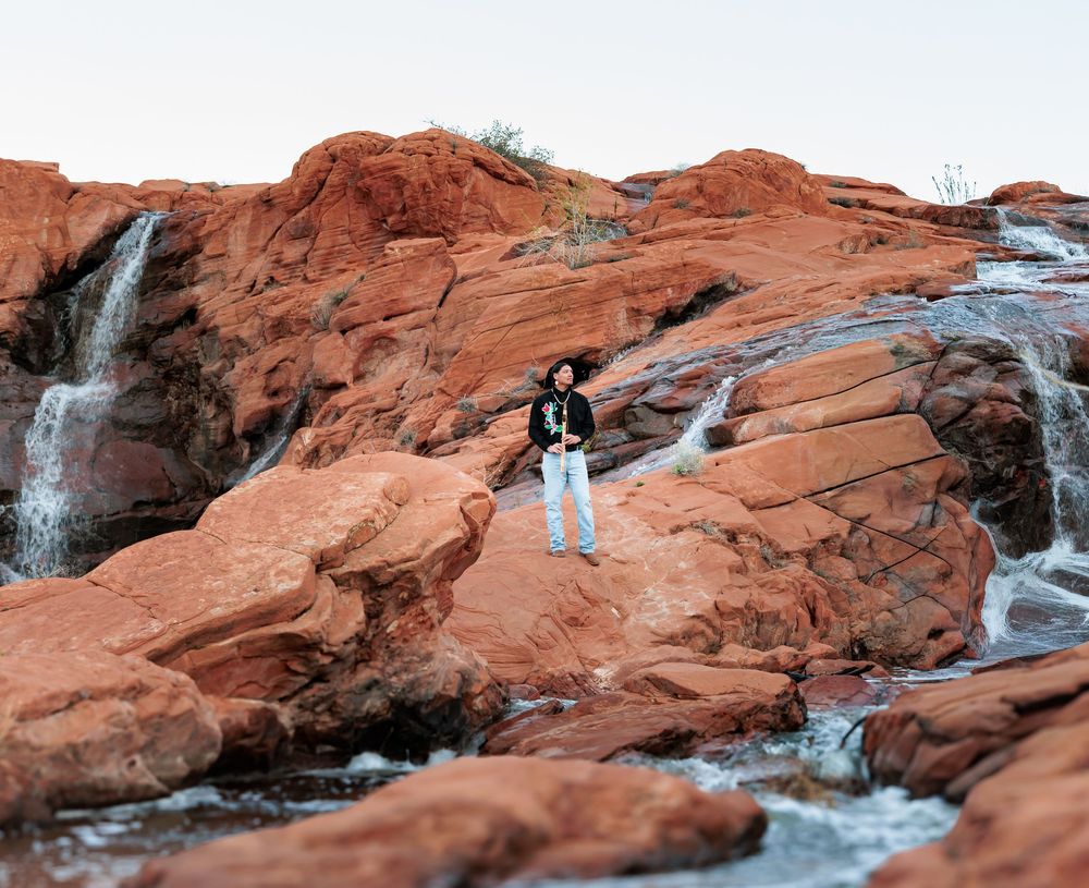 Person standing on red rock formations beside flowing waterfalls in a scenic desert canyon landscape.