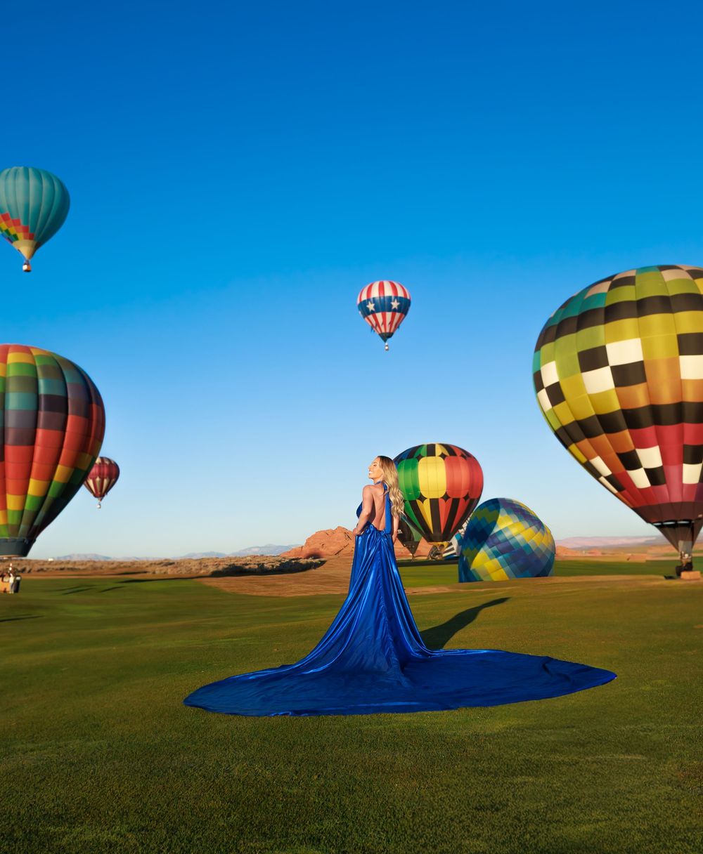 Woman in blue gown stands among colorful hot air balloons on green field under clear blue sky.