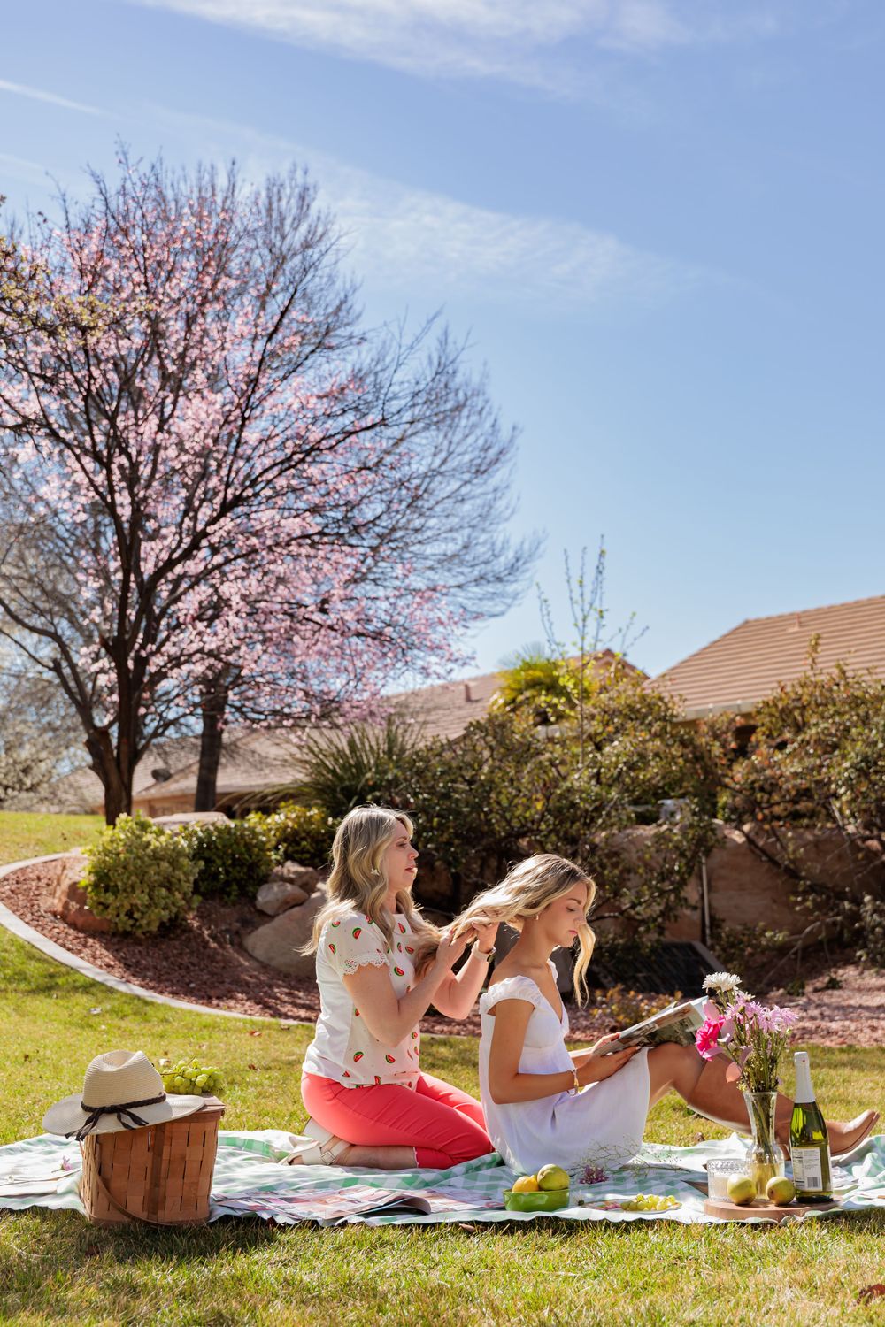 Two women enjoying a spring picnic on a blanket in a sunny garden with a blooming pink cherry tree.