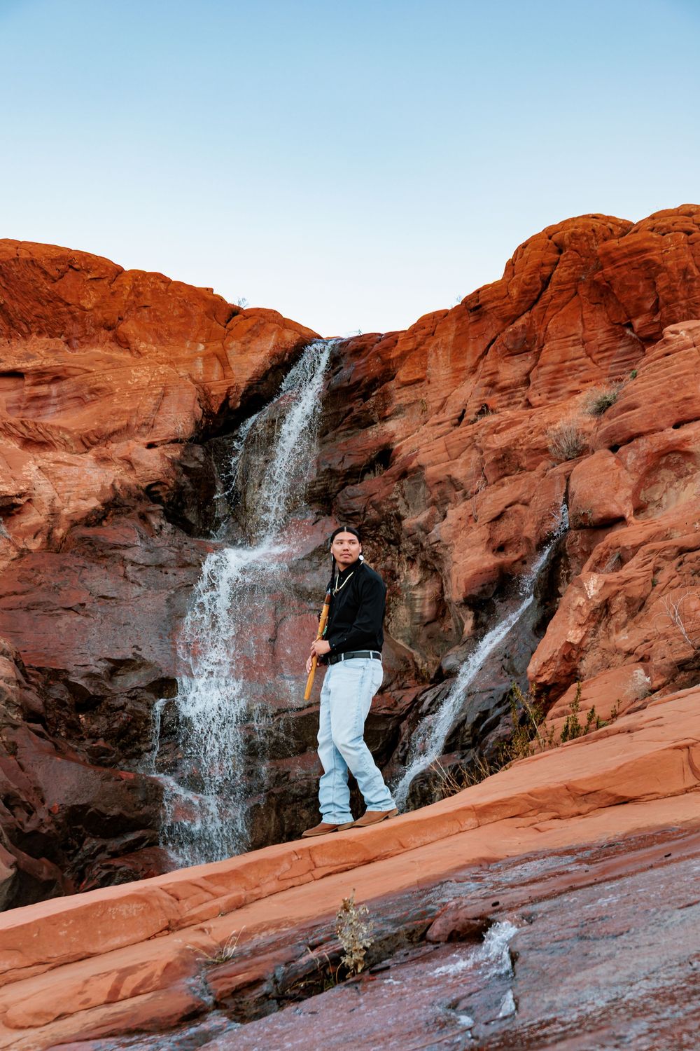 Person standing on red rock formations in front of a cascading waterfall in a desert canyon landscape.